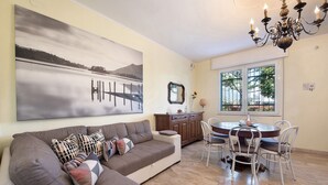 Uma sala de estar com sofá, mesa de jantar, cadeiras, um candelabro, um quadro e uma janela.