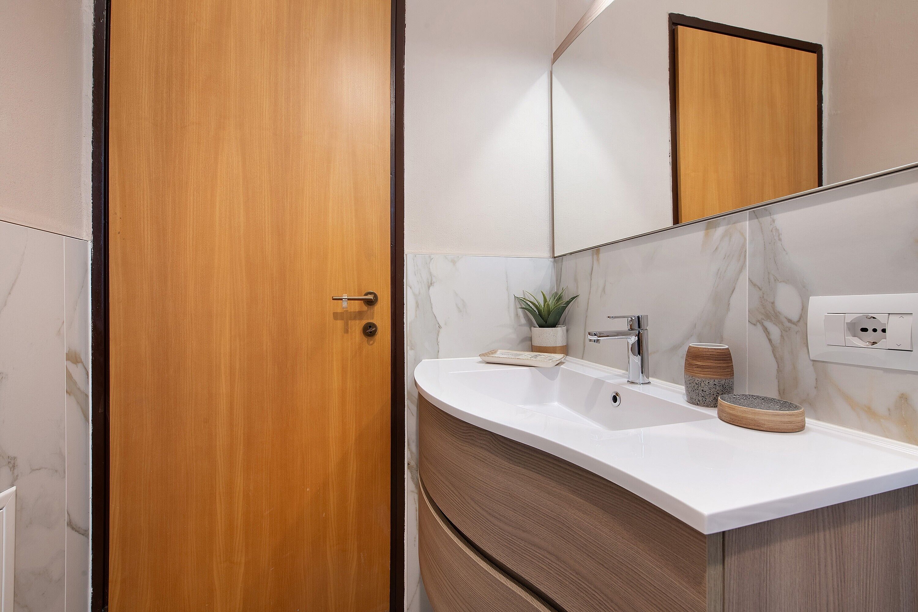 Une salle de bain avec une porte en bois, un lavabo blanc, un miroir et un mur en marbre.