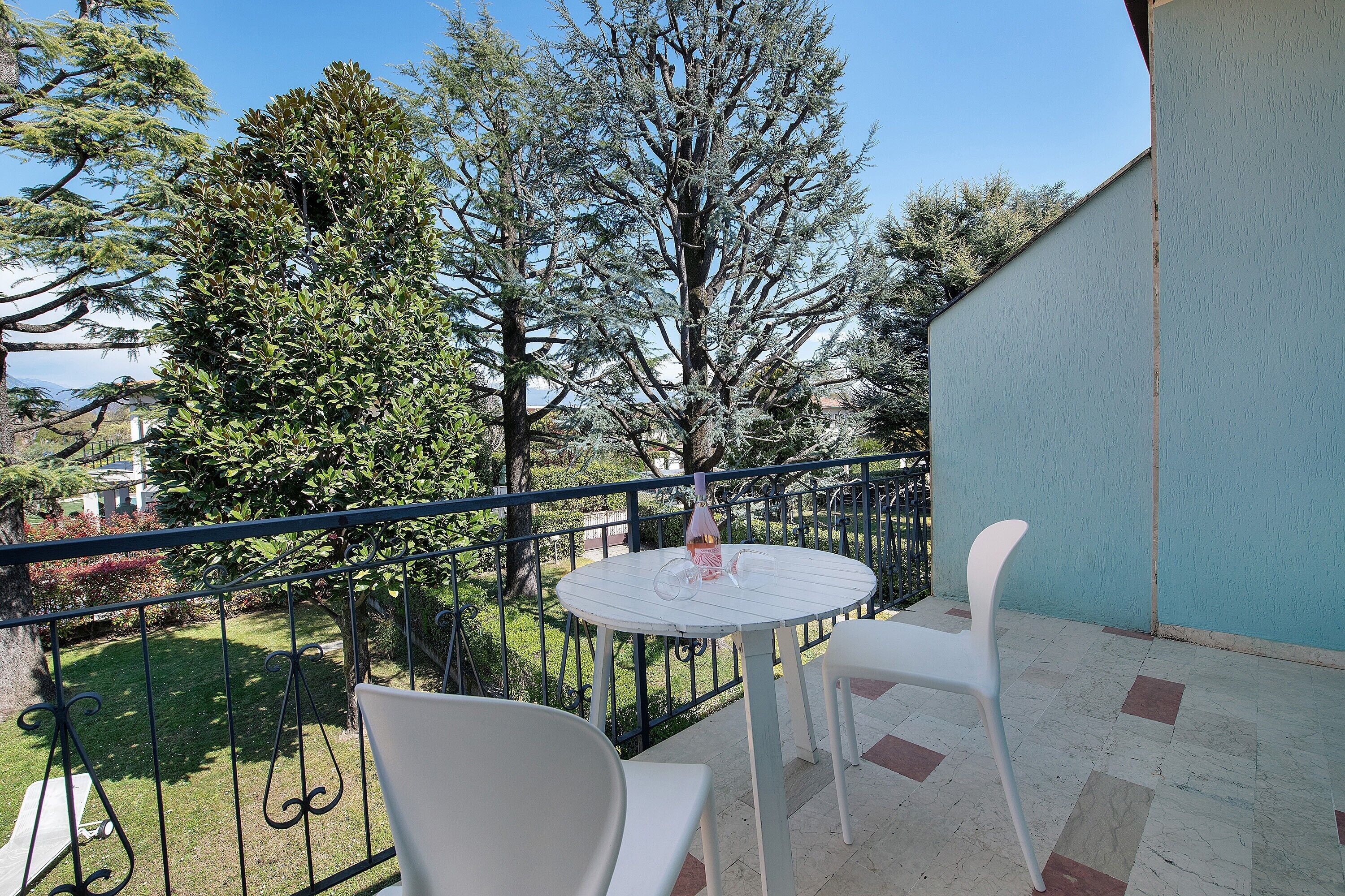Een balkon met een ronde tafel, twee stoelen en uitzicht op een boom en tuin.
