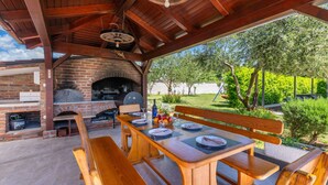 Een terras buiten met een bakoven, houten tafel, banken en uitzicht op een tuin.