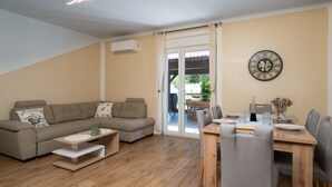Een woonkamer met een bank, salontafel, eettafel en stoelen. Er hangt een klok aan de muur en er staat een airconditioner.