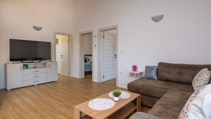 Een woonkamer met een bruine bank, een houten salontafel en een televisie op een witte kast. Er zijn twee deuren die naar andere kamers leiden.