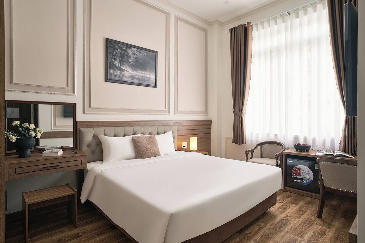 Hotellihuone, jossa on suuri sänky, yöpöytä lampun kanssa, pieni yöpöytä kulhon kanssa, tuoli, työpöytä tietokoneella ja televisio.