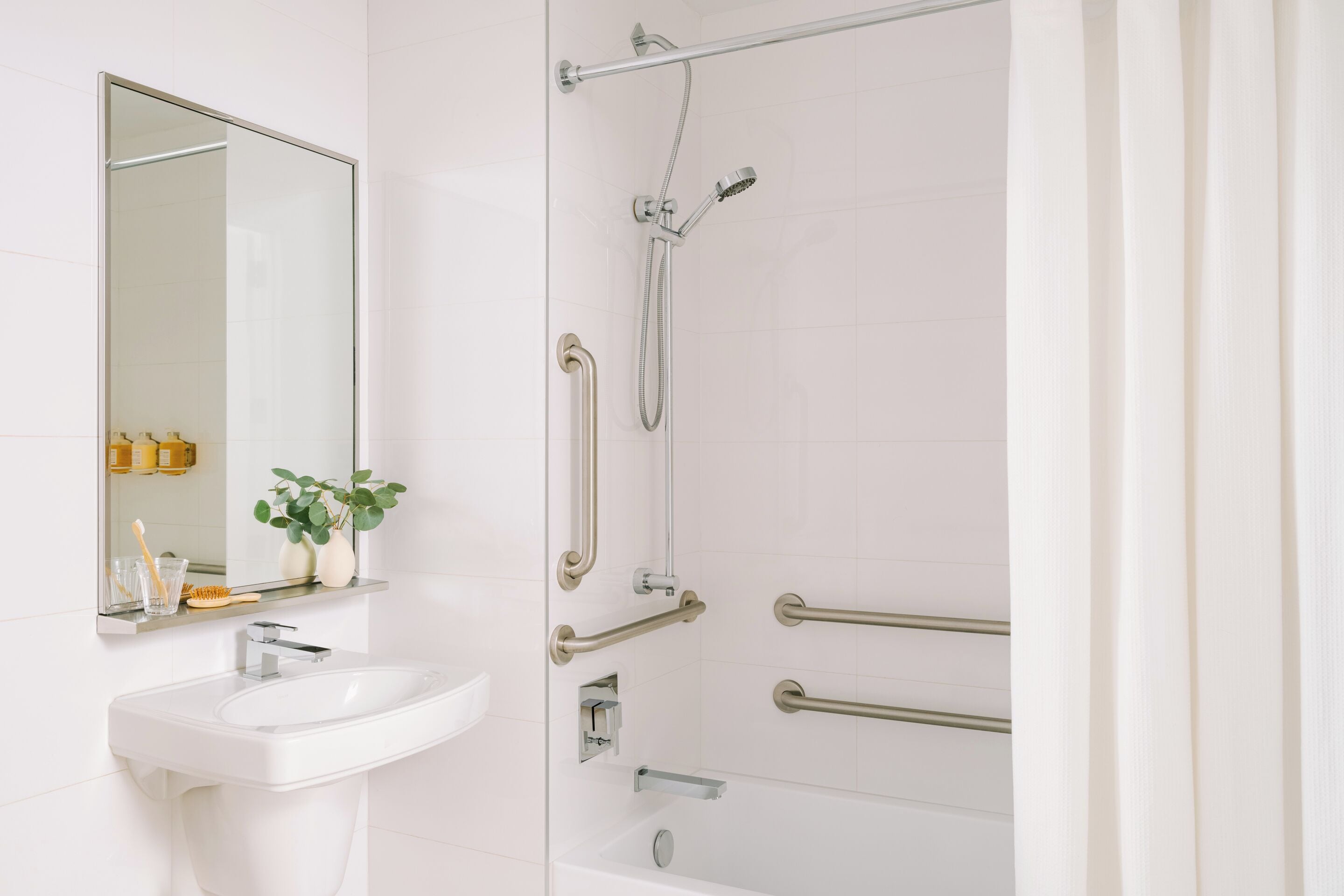 A bathroom with a white sink, mirror, and shower with grab bars.