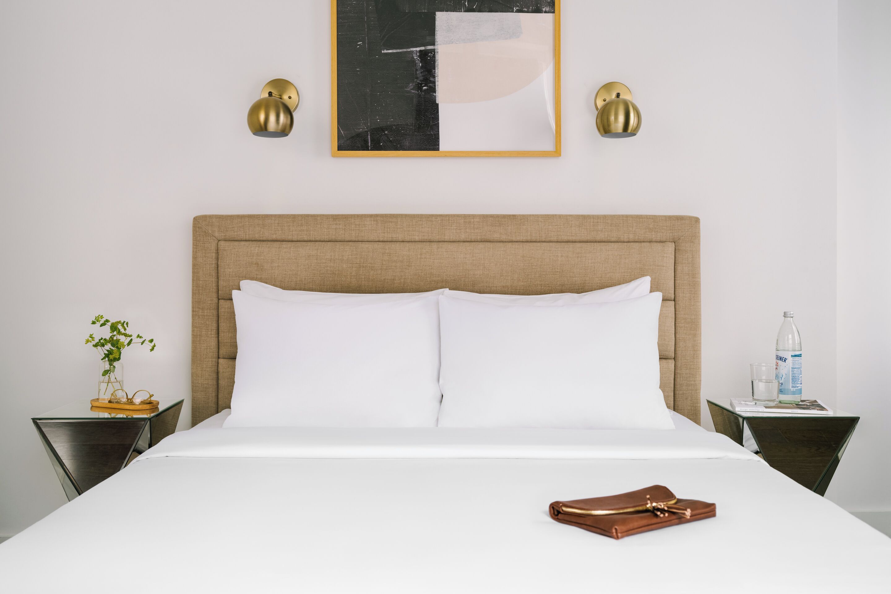 A neatly made bed with white linens, a brown headboard, and a brown handbag on the bed. There are two bedside tables, one with a plant and the other with a bottle and glass.