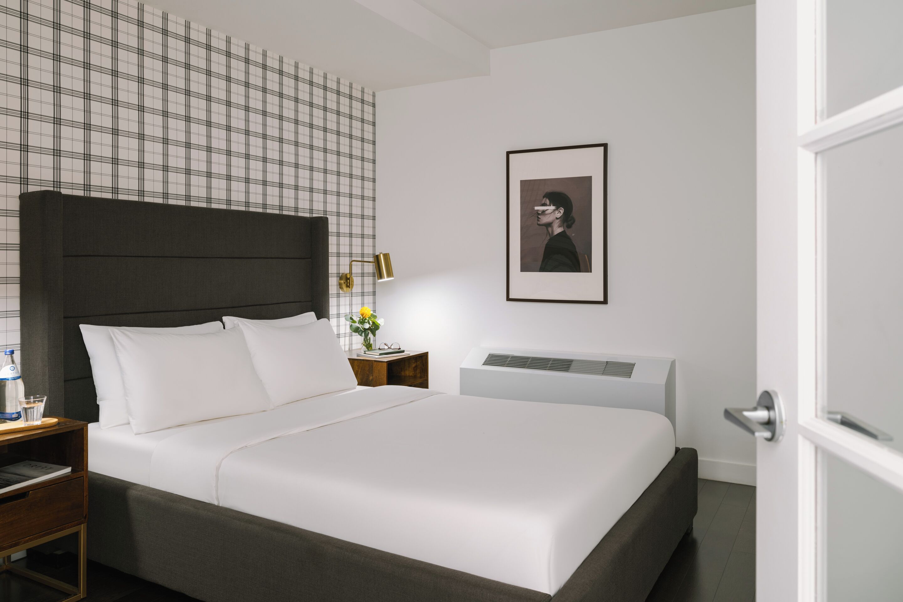 A modern hotel room with a large bed, a nightstand, a bedside table with a lamp, a framed picture on the wall, and a white door.