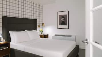 A modern hotel room with a large bed, a nightstand, a bedside table with a lamp, a framed picture on the wall, and a white door.
