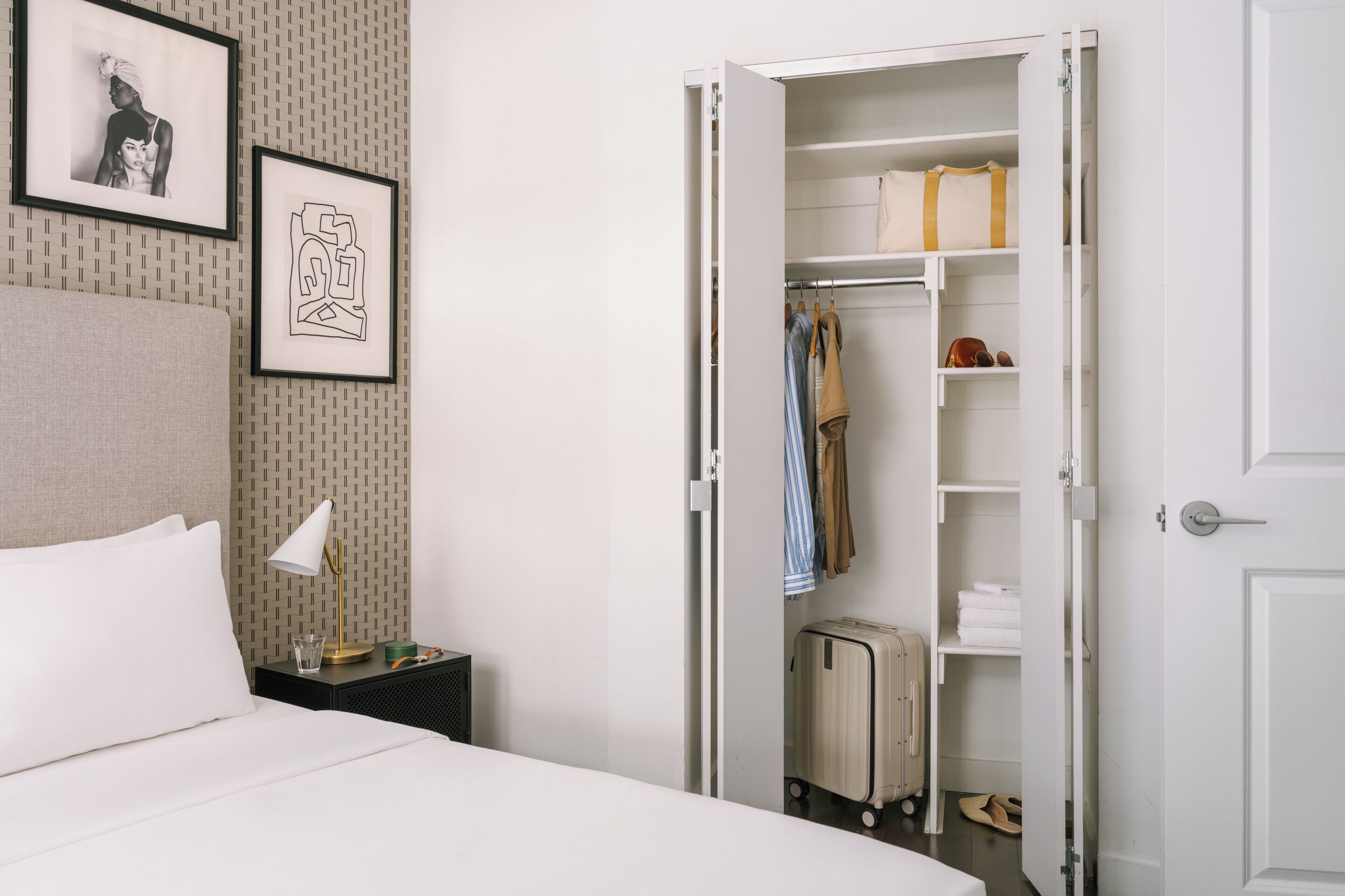 A neatly arranged hotel room with a bed, a bedside table with a lamp, a wardrobe, and a framed picture on the wall.