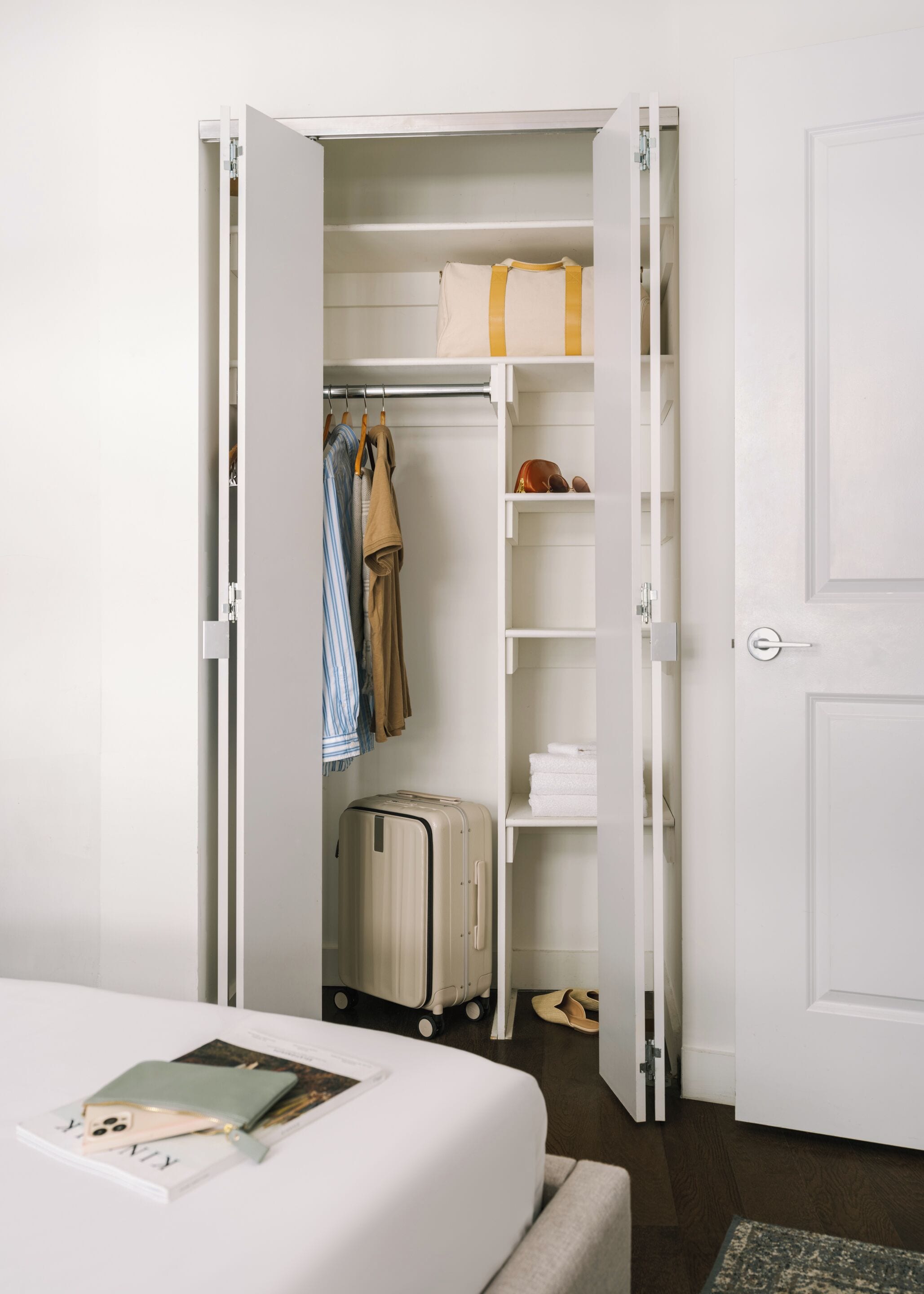 A room with a bed, a suitcase, a closet with hanging clothes, and shelves with folded towels.