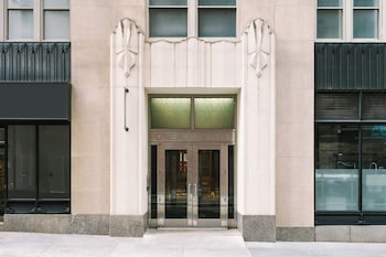 A neatly made bed with white linens, a headboard, a small bedside table with a lamp, and a window with a view of buildings. at Nester Finn Financial District