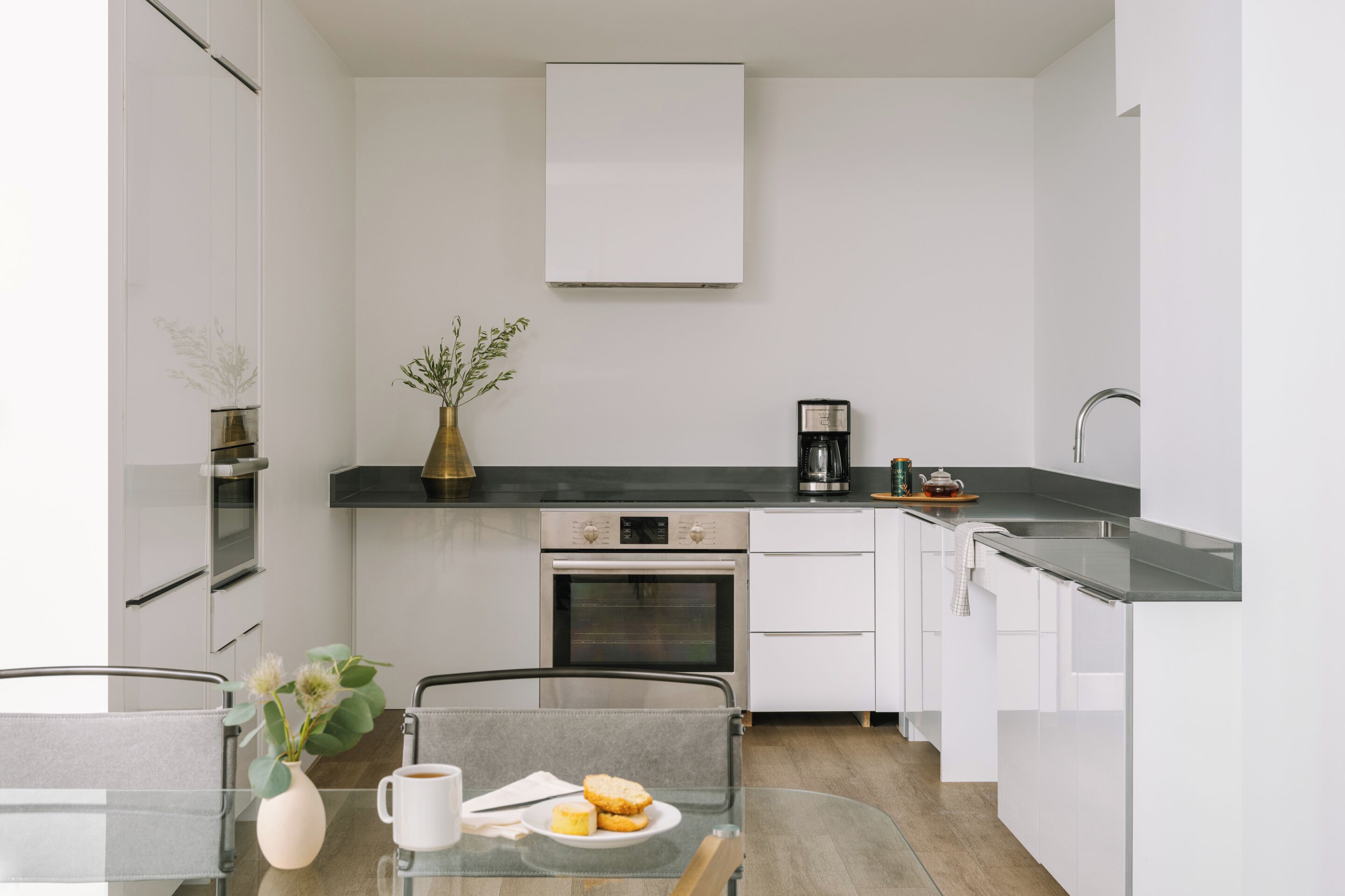 A modern kitchen with white cabinets, a black countertop, a built-in oven, and a dining area with a glass table and chairs.