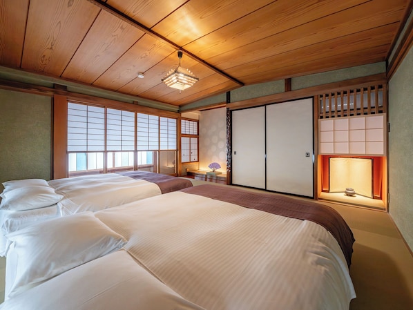 A traditional Japanese-style room with tatami flooring, a shoji screen, and a sliding door. There are two beds with white bedding and a small table with a lamp.