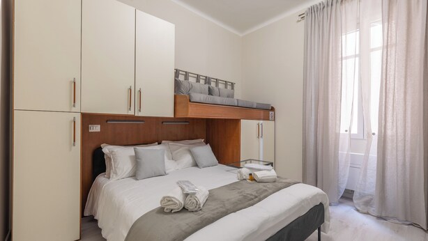Quarto com cama de solteiro e beliche, guarda-roupa espaçoso, mesa de cabeceira com abajur e janela com cortinas.