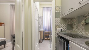 Um corredor estreito leva a um banheiro com cortina de chuveiro, pia pequena e vaso sanitário. No quarto adjacente, há uma cama com cabeceira e uma cadeira.