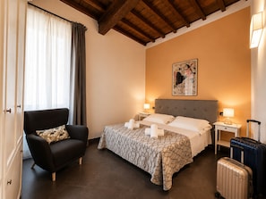 Ein Hotelzimmer mit einem Bett, einem Sessel, einem Nachttisch mit Lampe, einem Wandbild und einem Koffer.