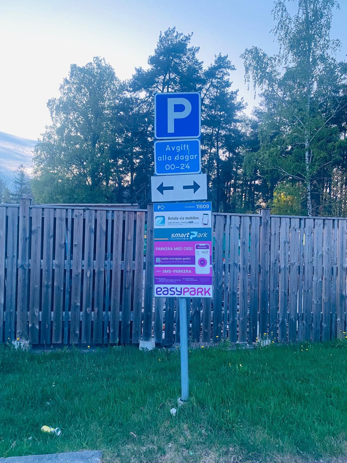 A parking sign with a blue 'P' and directional arrows, a sign for 'Avgift alla dagar 00-24', and a 'smartpark' advertisement.