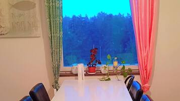 A dining area with a white table, black chairs, a window with curtains, and a view of trees.