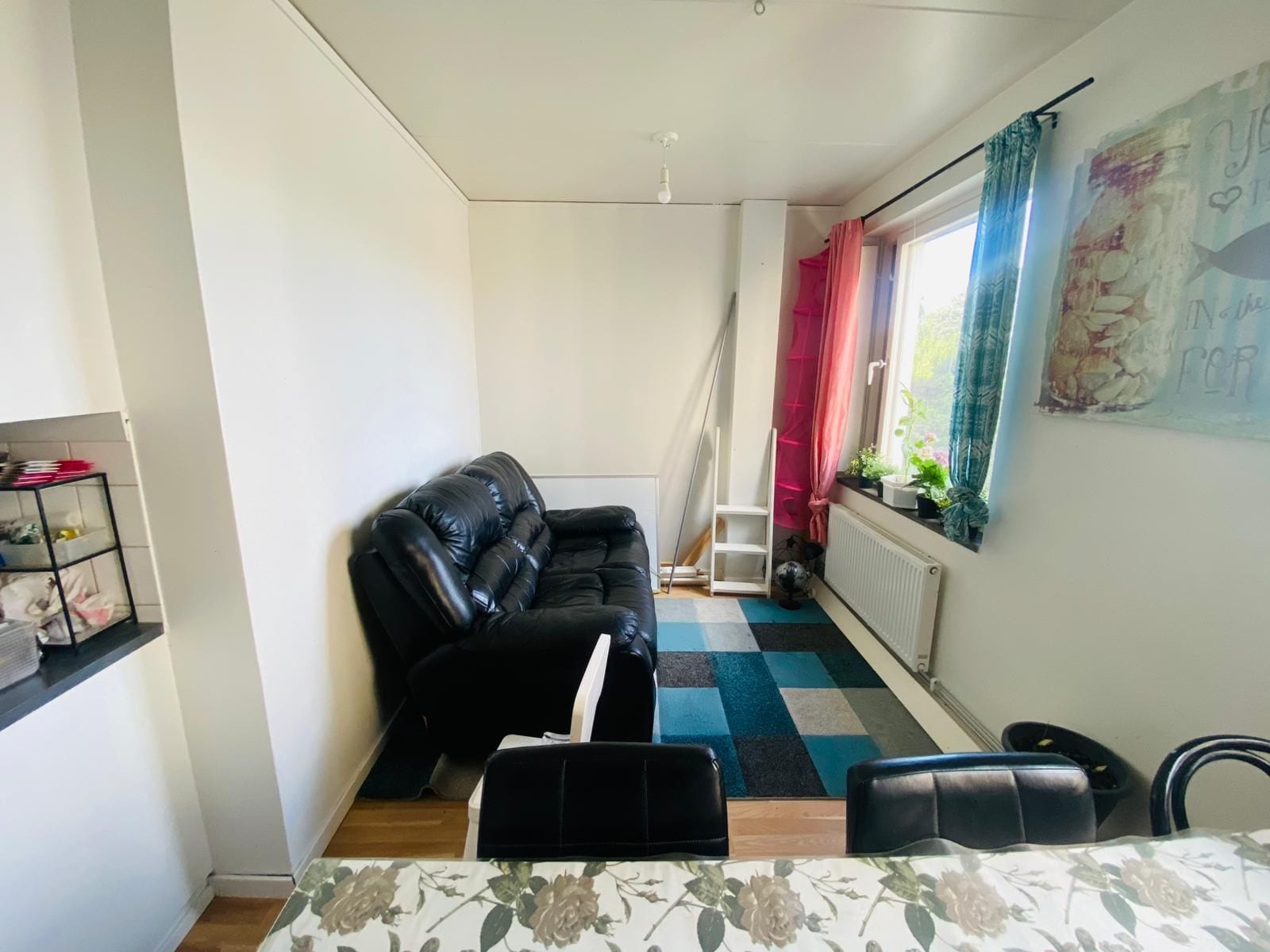 Ein kompakter Wohnbereich mit einer schwarzen Couch, einem kleinen Esstisch und einem Heizkörper in der Nähe des Fensters.