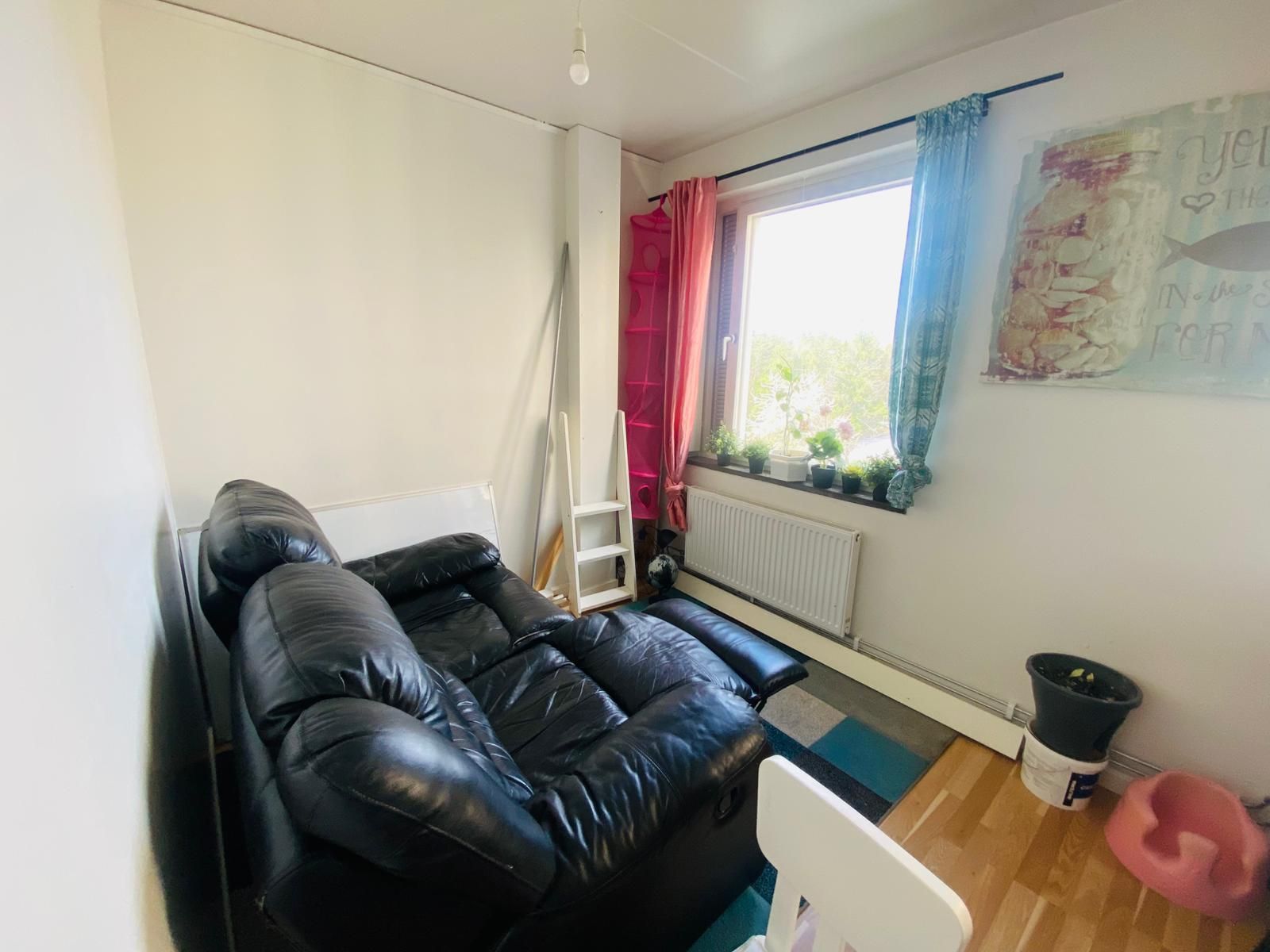 A compact living space with a black leather sofa, a radiator, a window with curtains, and a small potted plant on the windowsill.