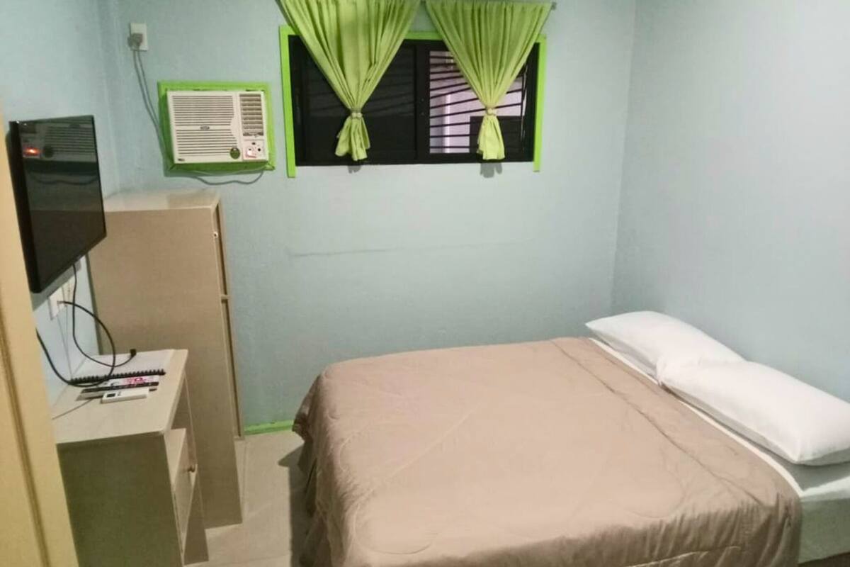 Ein Schlafzimmer mit einem Bett, einem Schreibtisch mit Computer, einem Fernseher und einem Fenster mit Vorhängen.