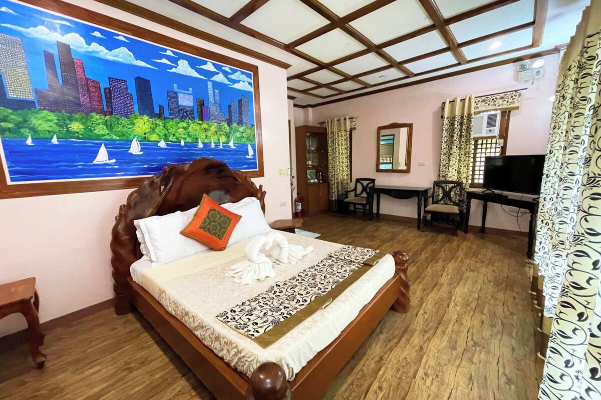 Una cama bien tendida con un cuadro de paisaje urbano, cabecera de madera y colcha estampada.