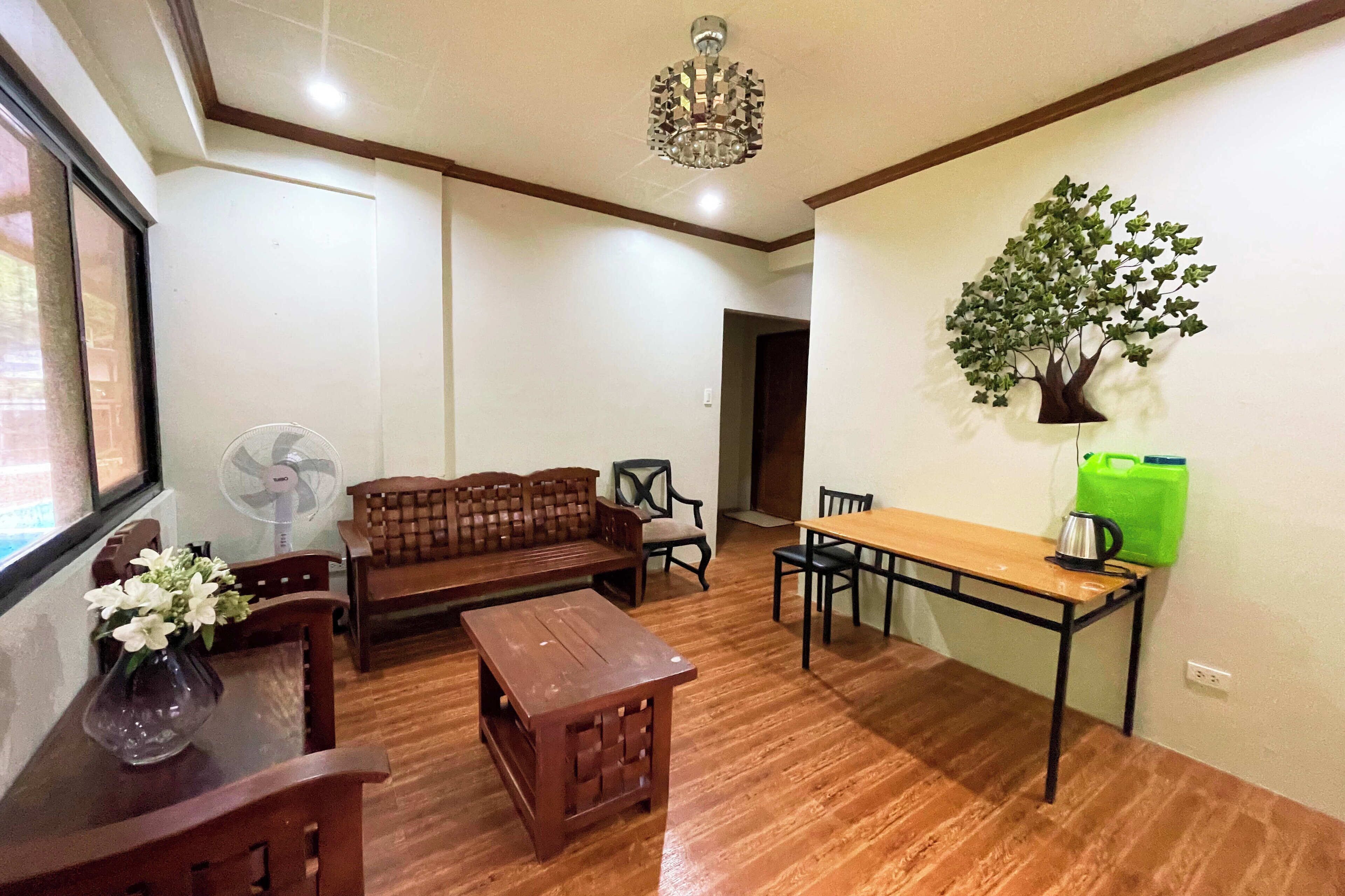 Una sala de estar con piso de madera, sofá de madera, mesa de centro de madera, una mesa de comedor con sillas, un ventilador y una araña.
