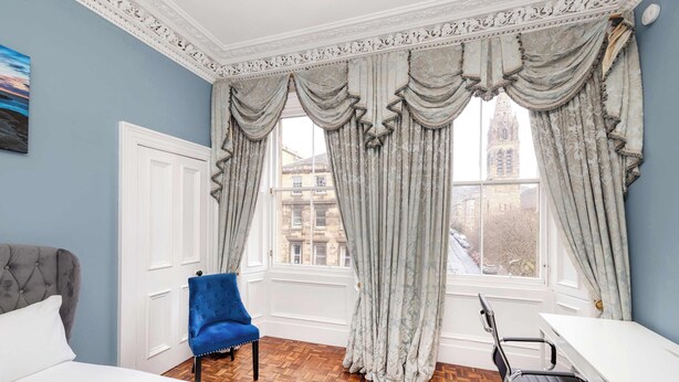 A hotel room with a bed, a blue armchair, a desk, and large windows with curtains.