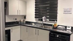 A modern kitchen with white cabinets, a black countertop, a built-in dishwasher, and a window with blinds. - Contractor/Corporate-Friendly Home | Sleeps 6 | Long Stays (Bolton)