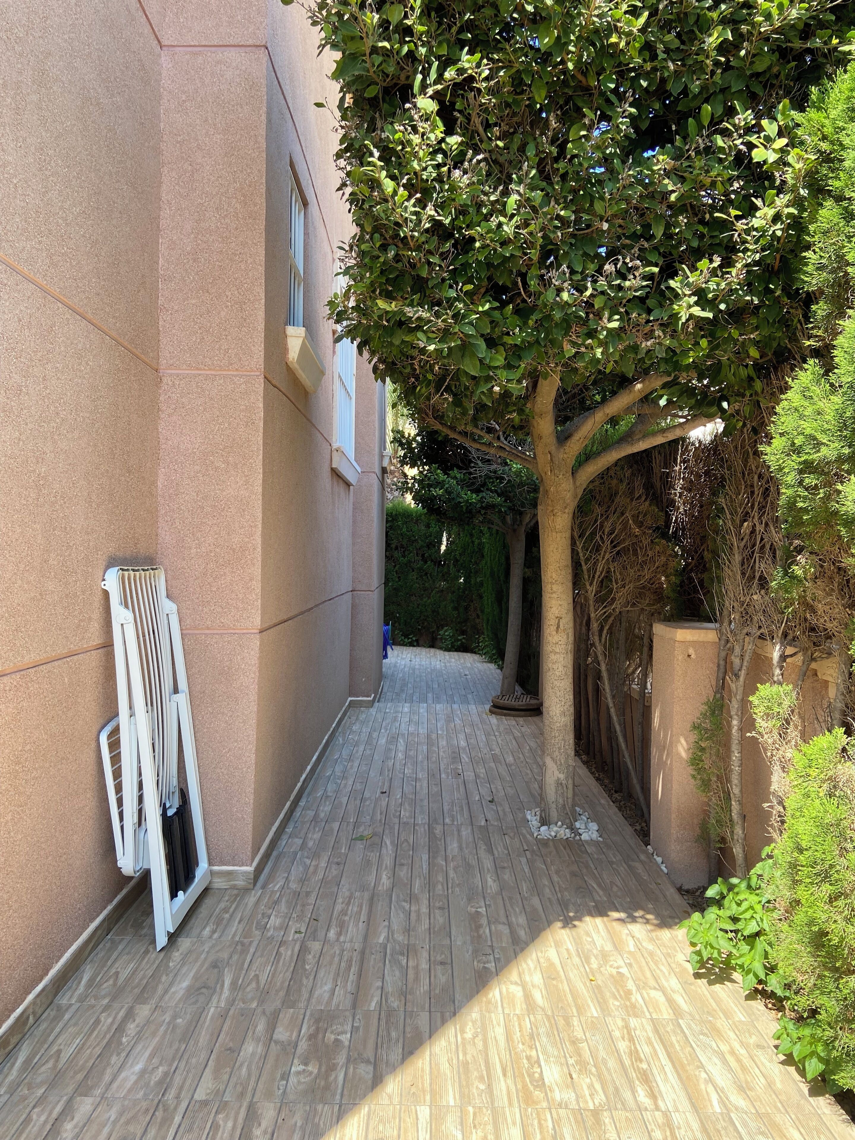 A narrow wooden pathway flanked by a building with a beige exterior and a tree providing shade.