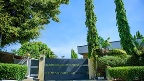 Una entrada con reja y un edificio moderno, rodeado de abundante vegetación y altos árboles.