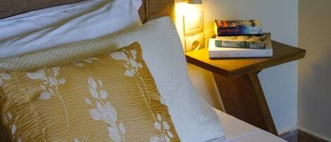 A neatly made bed with floral-patterned pillows, a bedside table with a lamp, and a stack of books.