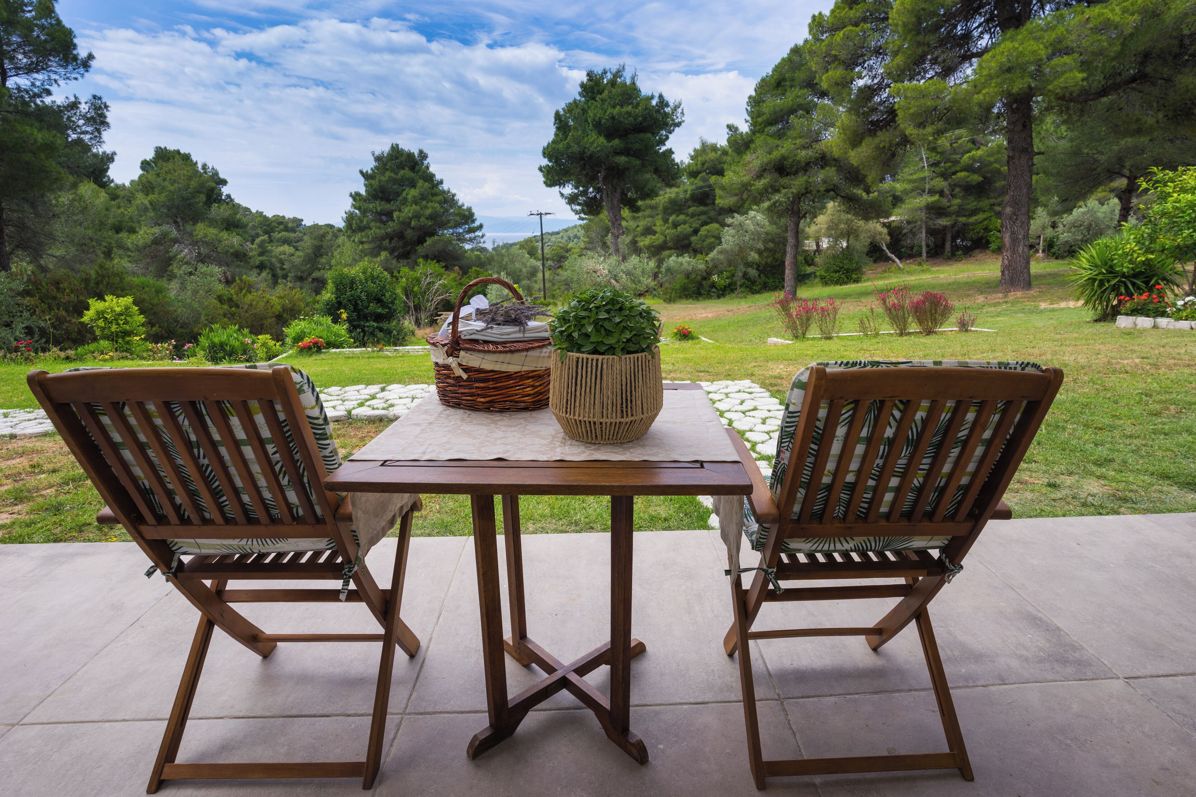 An outdoor patio with two wooden chairs and a table, surrounded by a garden with various plants and trees.