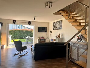Un salon moderne avec un escalier, un téléviseur et un coin repas visible à travers une porte vitrée.