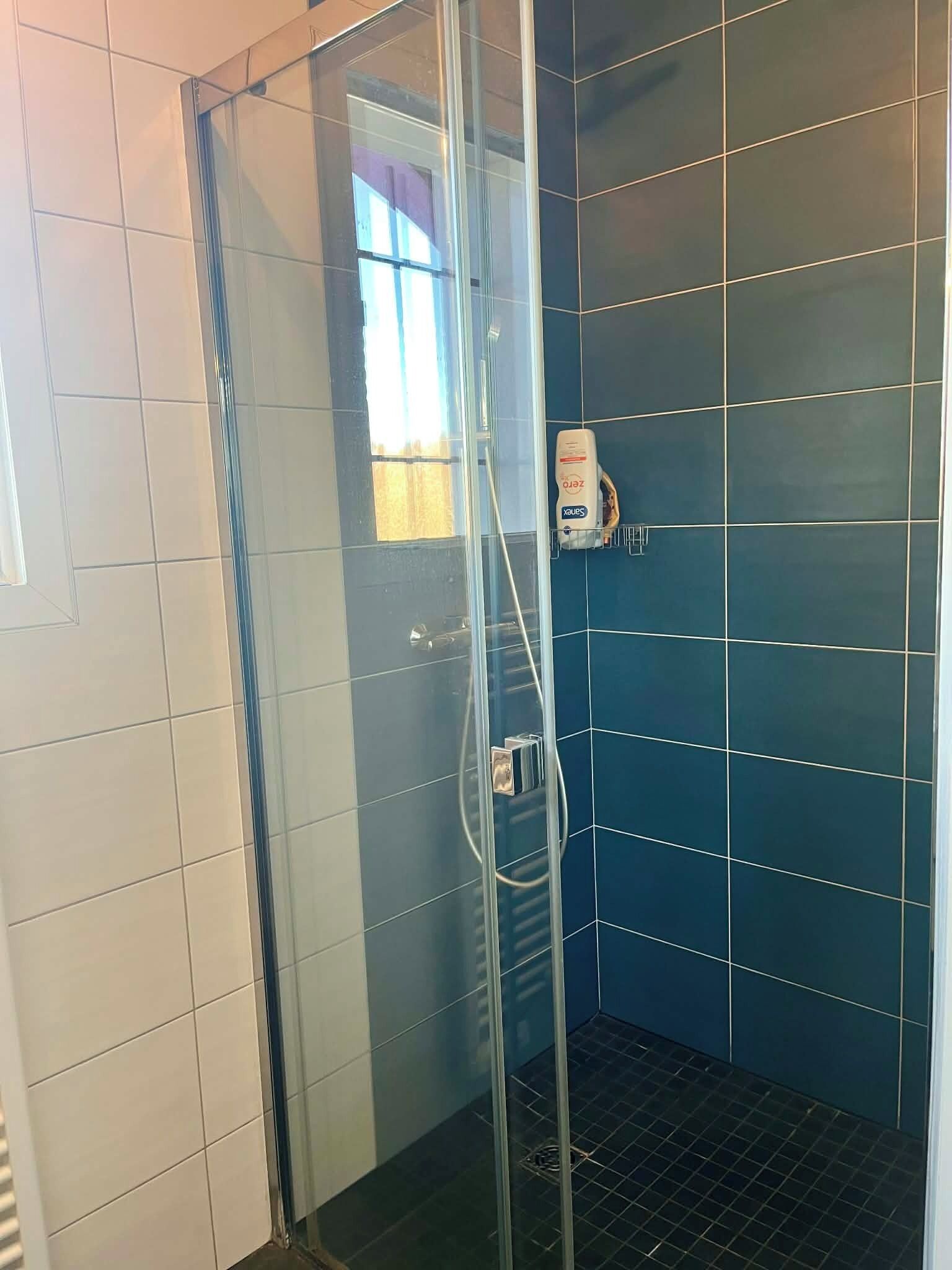Una ducha con puerta de vidrio, una ventana con cortinas y un dispensador de jabón en la pared.