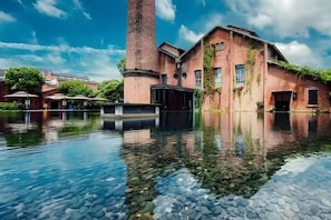 Een historisch gebouw met een grote schoorsteen, een reflecterend waterpartij ervoor en een heldere lucht.