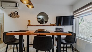 A dining area with a wooden table, black chairs, a TV mounted on the wall, and a window with a view of trees.