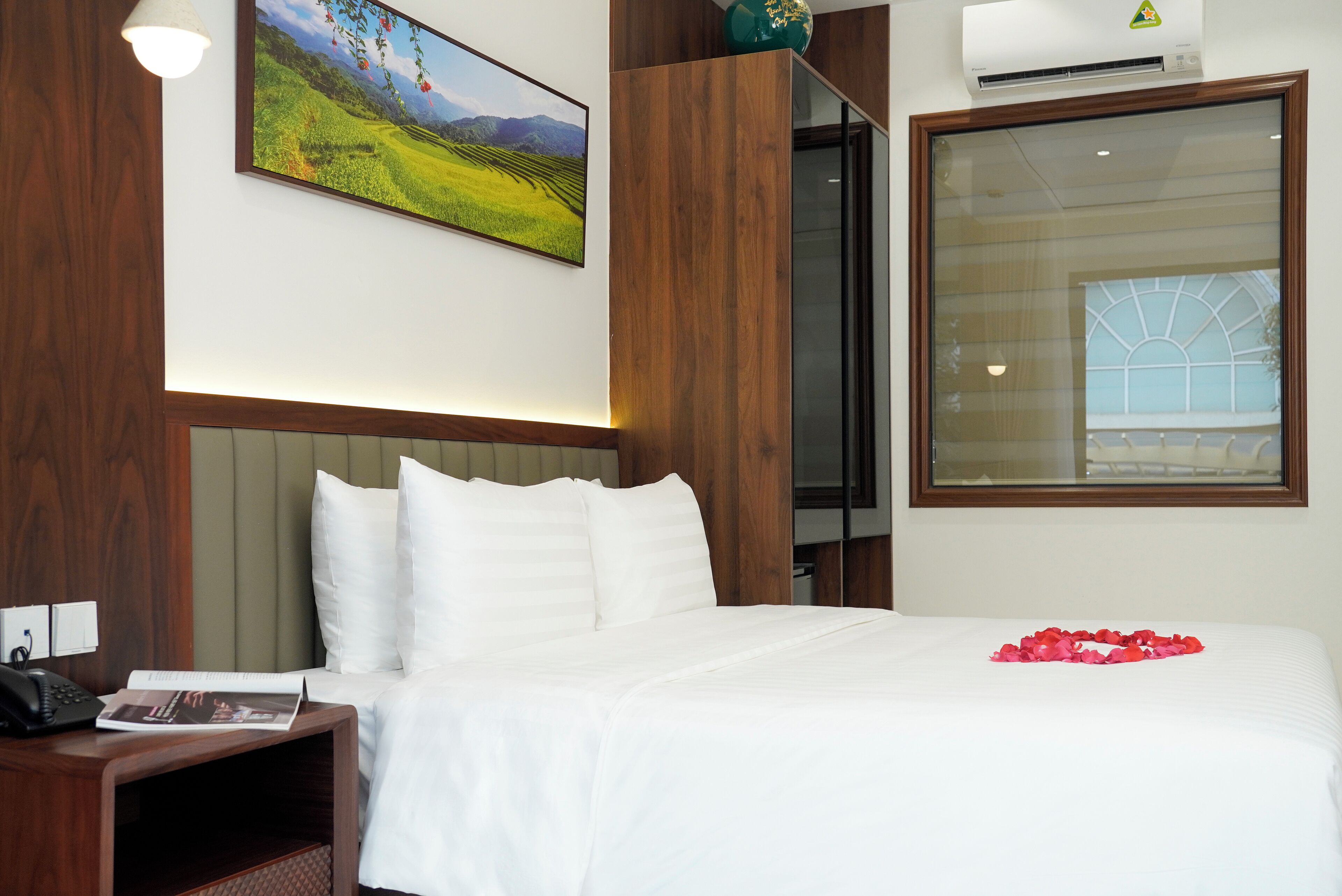 Habitación de hotel con una cama grande, cabecera de madera, mesita de noche con teléfono, un cuadro en la pared y ventana con vista a zonas verdes.