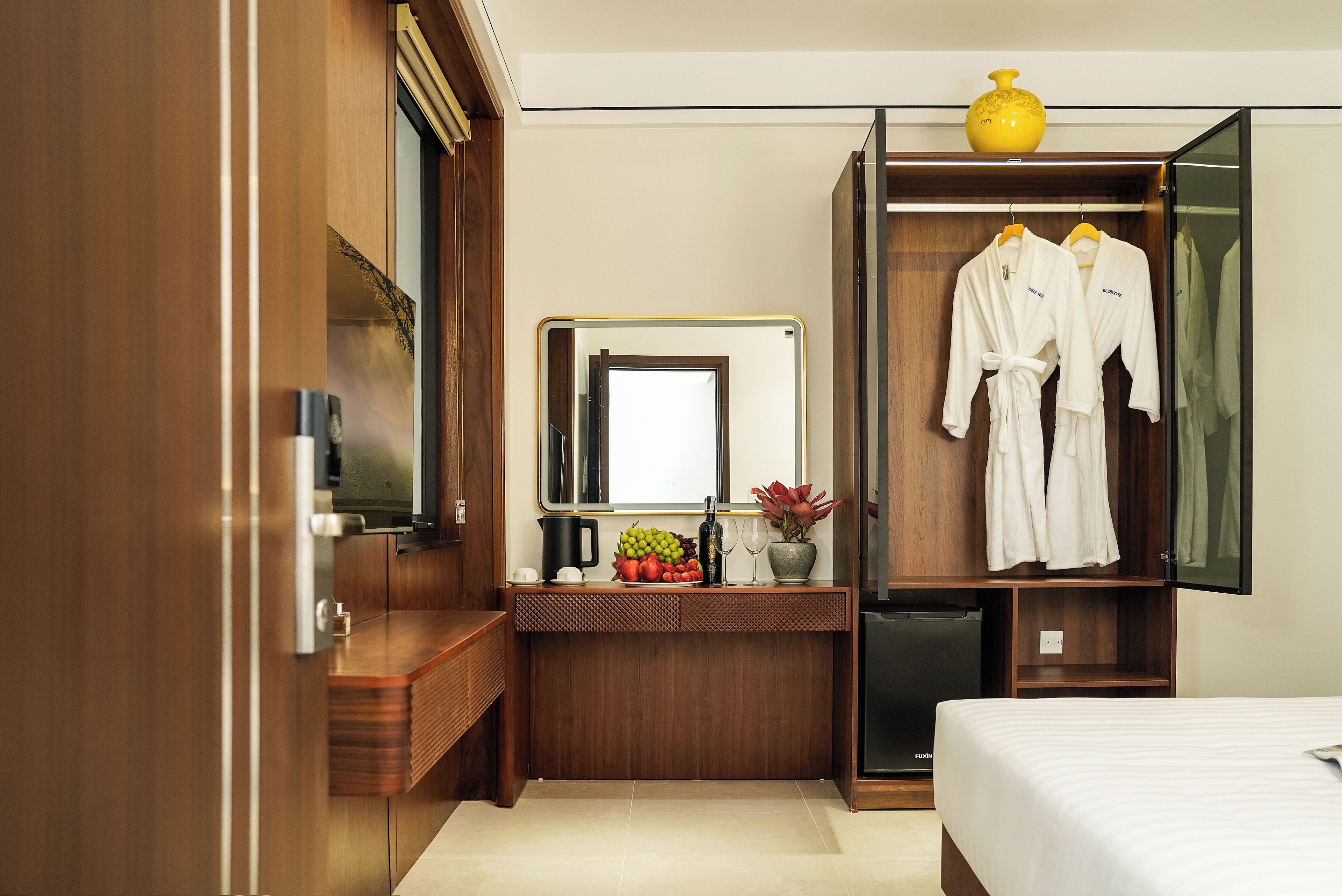 A modern hotel room with a wooden vanity, a mirror, a bed, and a wardrobe with hanging robes.
