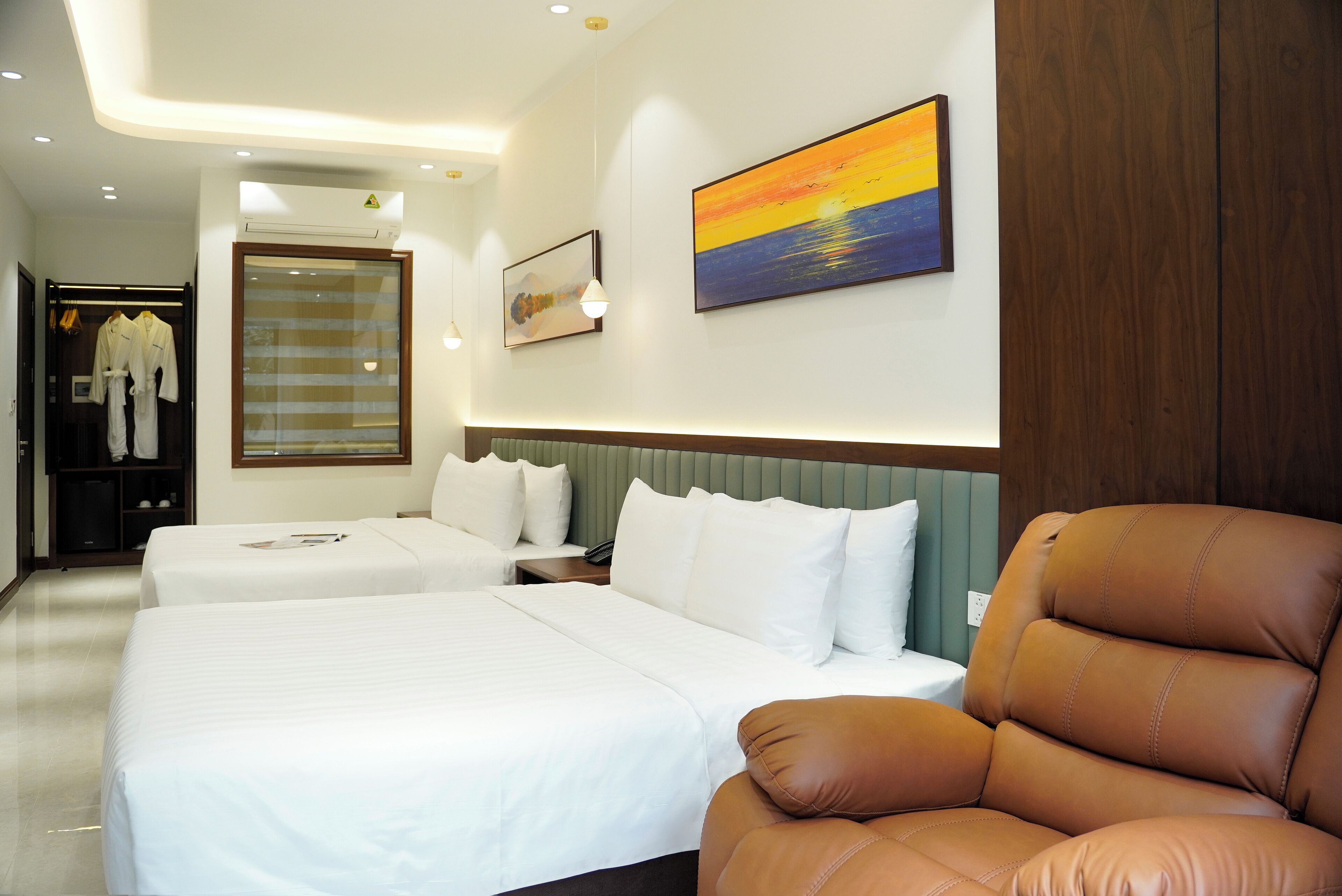 Habitación de hotel con dos camas, una butaca de cuero marrón, un cuadro de un atardecer y un armario con ropa colgada.