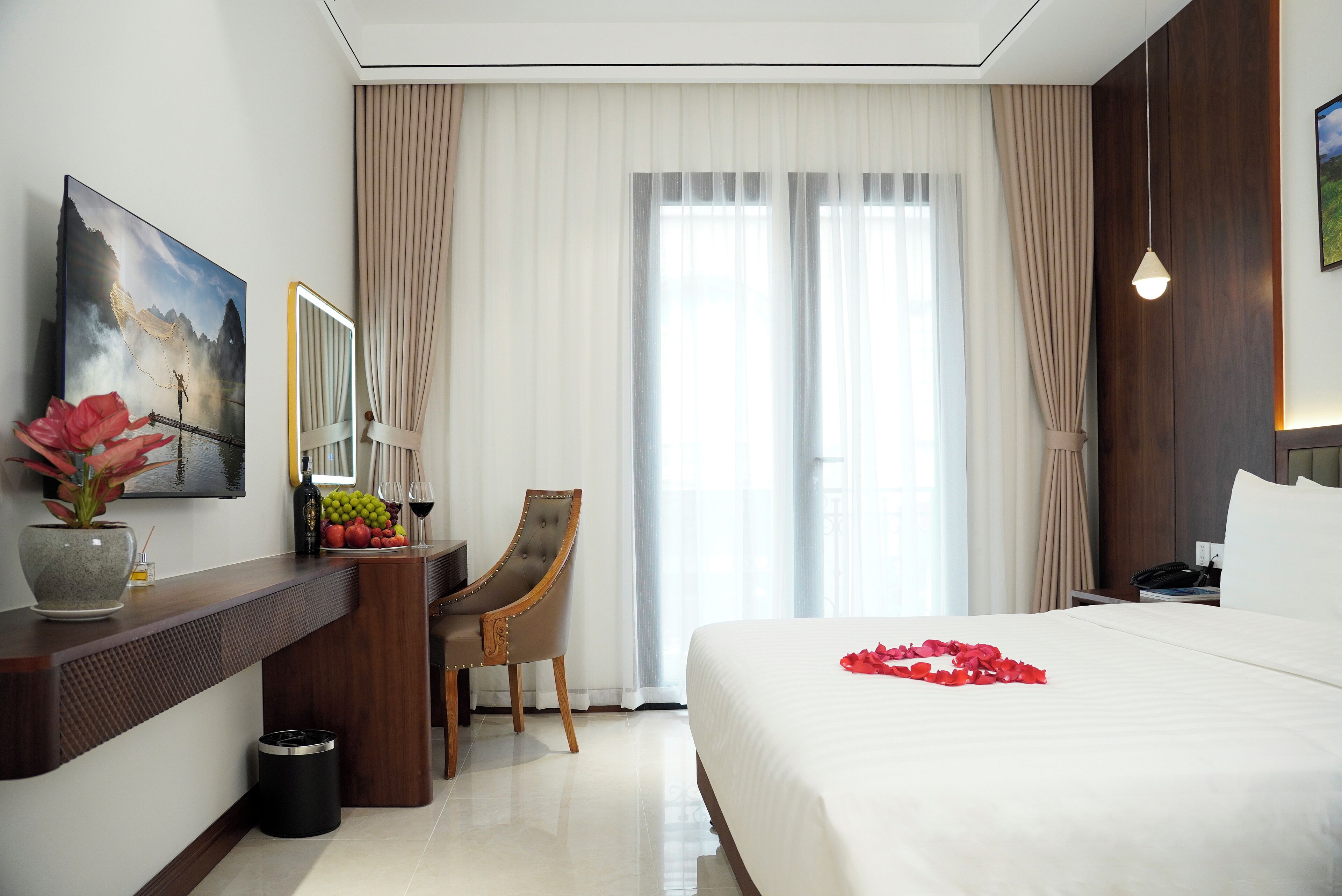 Ein Hotelzimmer mit Bett, Schreibtisch, Sessel, einem großen Fenster mit Vorhängen, einem Bild an der Wand und einer Vase mit Blumen.
