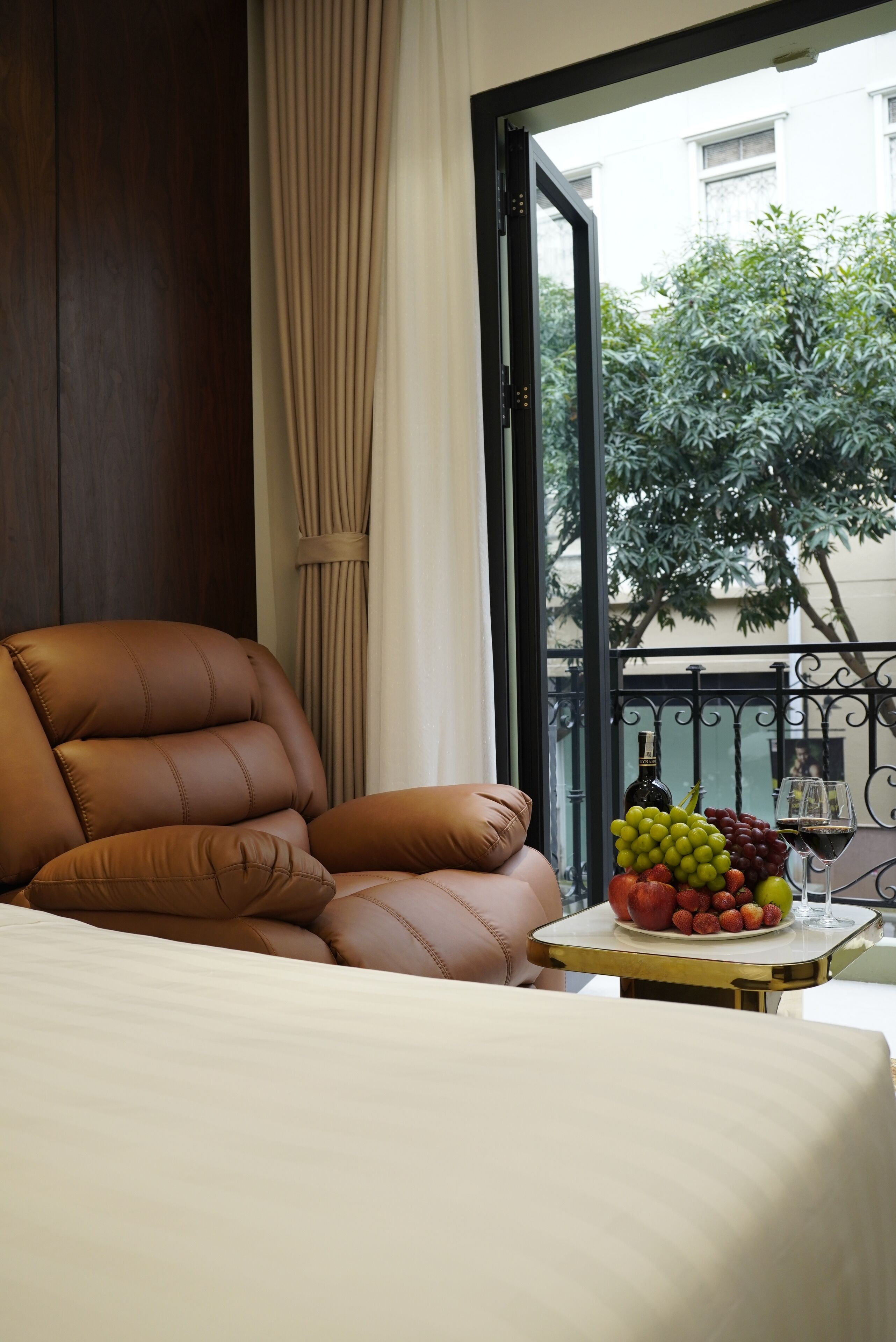 A hotel room with a leather armchair, a bedside table with a fruit bowl and wine glasses, a window with a view of a tree and buildings, and a curtain.