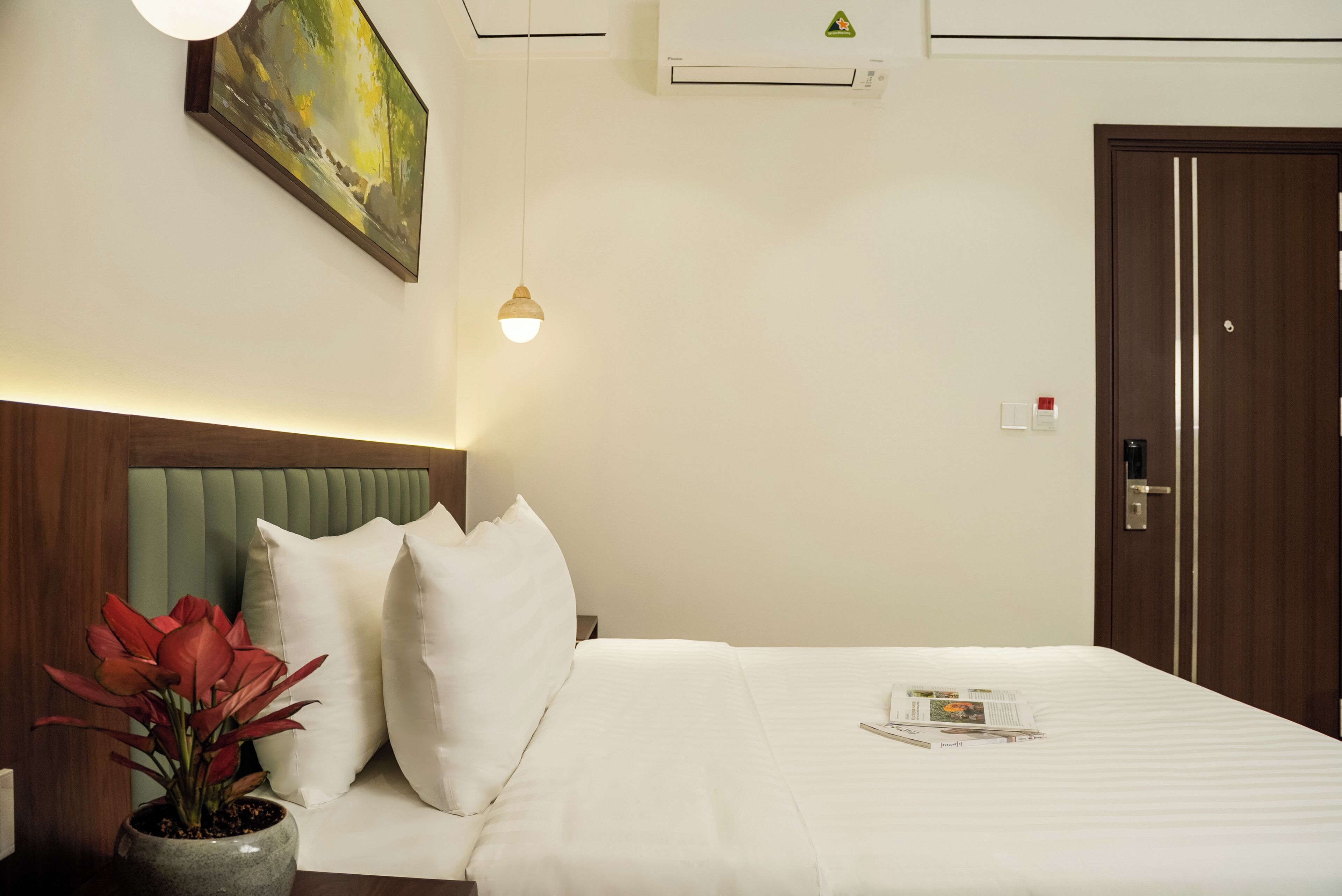 Une chambre d’hôtel avec un lit, des oreillers blancs, une plante en pot, un tableau au mur et une porte.