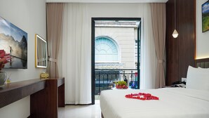 Habitación de hotel con una cama grande, un escritorio con espejo, vistas al exterior a través de una ventana y un balcón con barandilla.