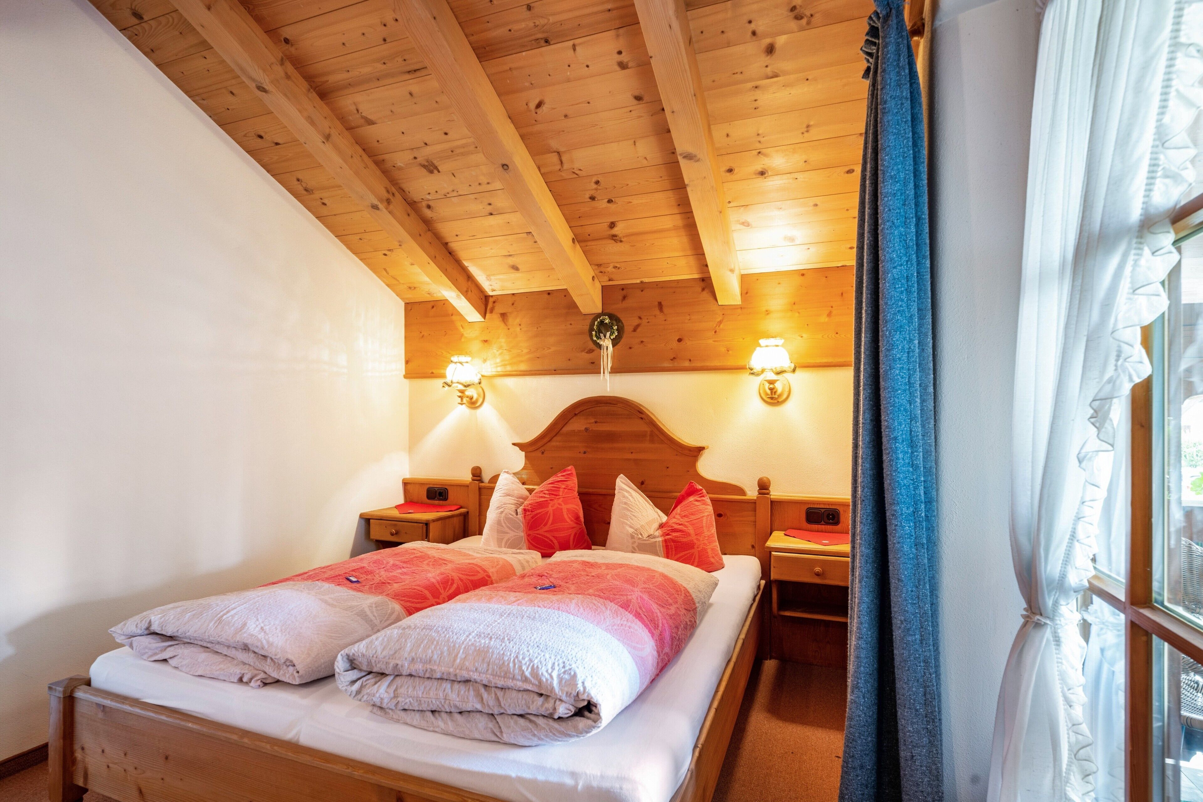 Ein Zimmer mit Holzbalken, einem Bett, zwei Nachttischlampen und einem Fenster mit Vorhängen.