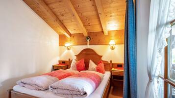 Ein Zimmer mit Holzbalken, einem Bett, zwei Nachttischlampen und einem Fenster mit Vorhängen.