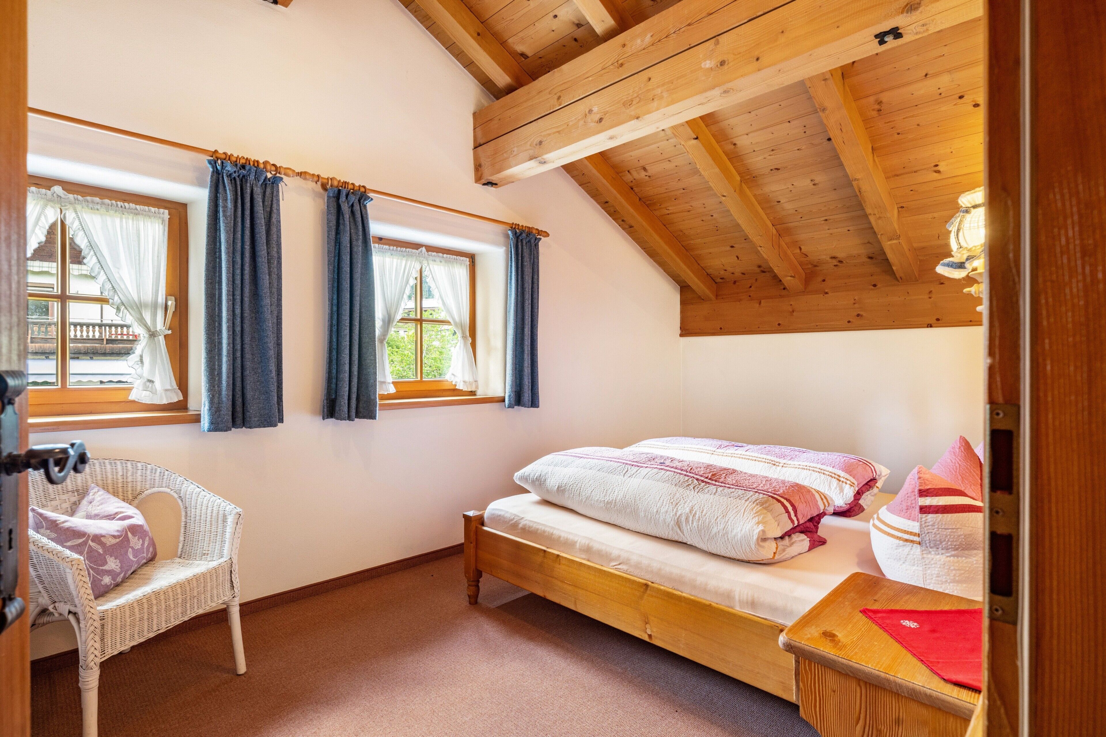 Un dormitorio con techo de madera, una cama individual, una silla y un ventanal con cortinas.