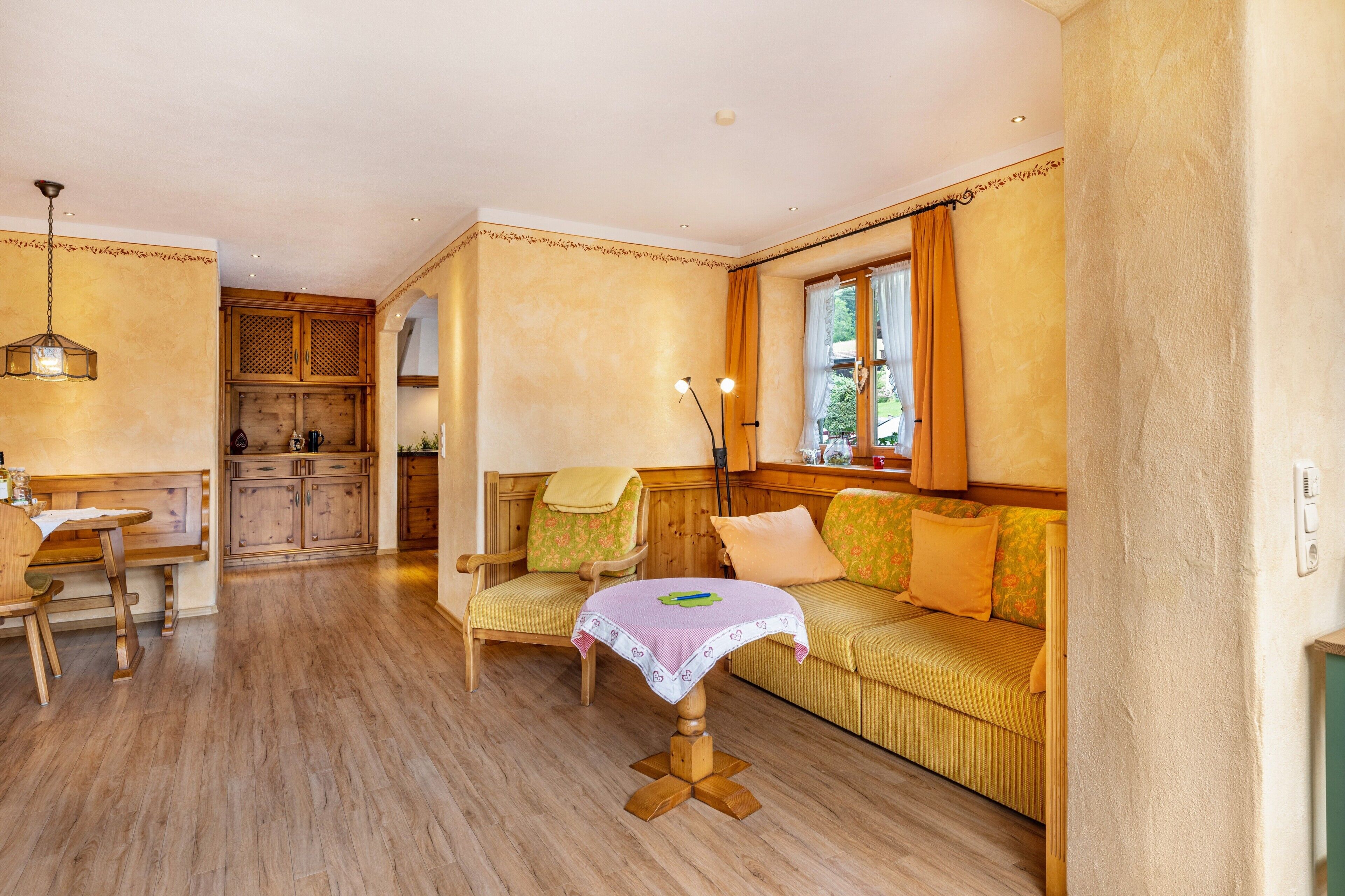 A cozy room with a wooden dining table, a sofa with cushions, a small table with a tablecloth, and a window with curtains.
