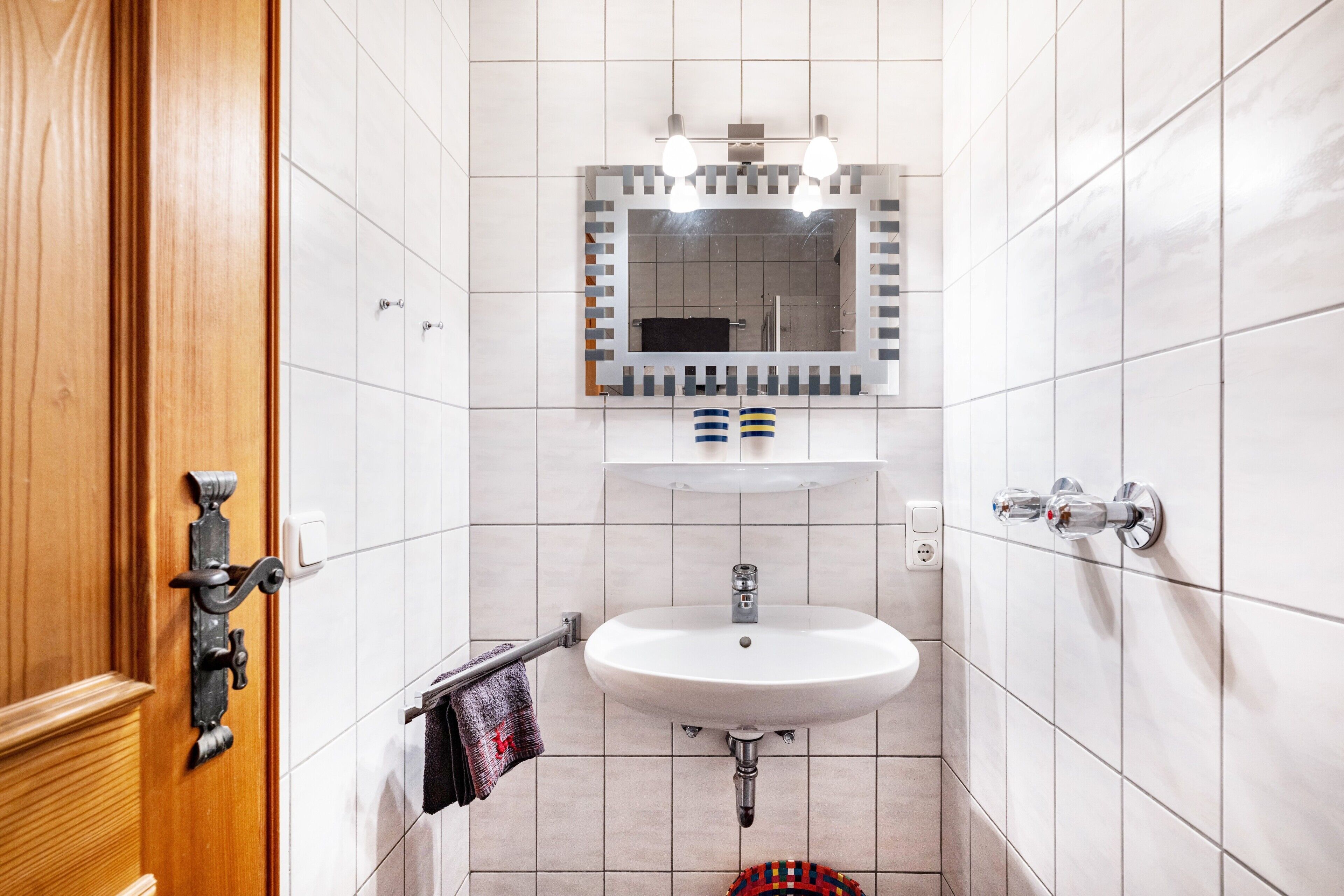 A bathroom with a wooden door, white tiled walls, a white sink, a mirror with lights, and a towel rack.