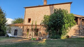 Un bâtiment en pierre avec un toit de chaume, une petite fenêtre et une cheminée. On aperçoit un fauteuil de jardin blanc et un arbre au premier plan.