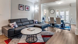 En moderne stue med en hjørnesofa, et spiseområde med bord og stole, og et synligt ur på væggen.